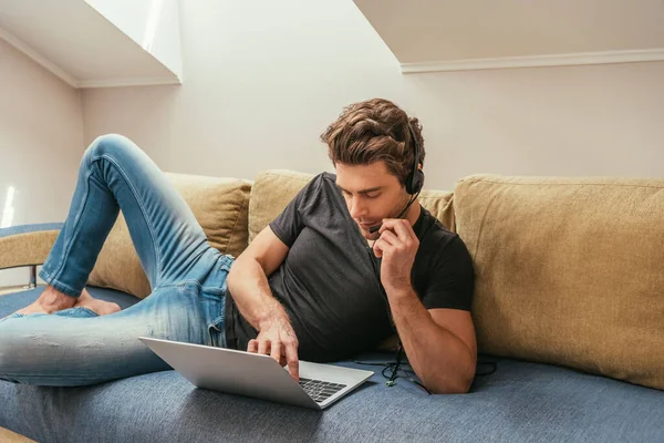 Bel homme dans le casque couché sur le canapé près d'un ordinateur portable tout en travaillant à la maison dans la chambre mansardée — Photo de stock