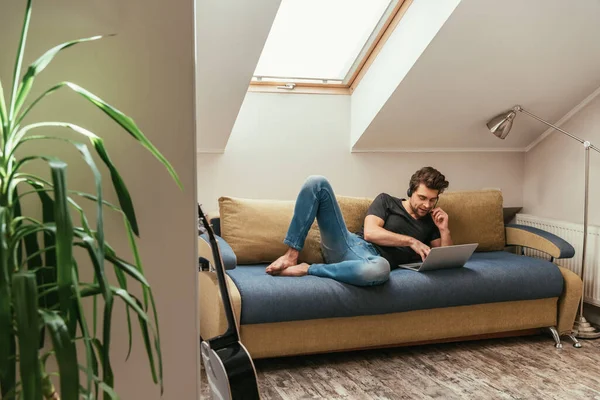 Hombre guapo en auriculares tumbado en el sofá cerca de la computadora portátil mientras trabajaba en casa en la habitación del ático - foto de stock