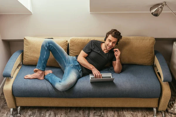Высокий угол обзора улыбающегося человека в гарнитуре, лежащего на диване рядом с ноутбуком и смотрящего на камеру — стоковое фото