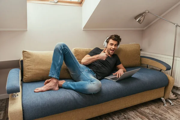 Excited man in wireless headphones showing thumb up while lying on sofa near laptop — Stock Photo