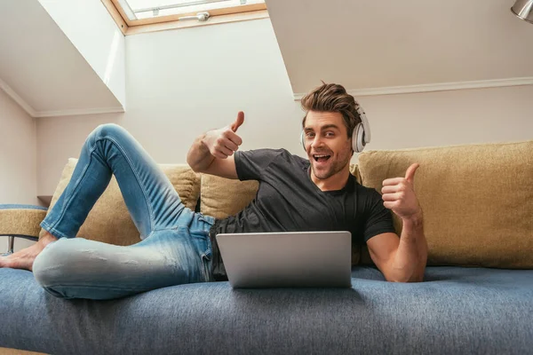 Homme heureux dans les écouteurs sans fil montrant pouces levés tout en étant couché sur le canapé près d'un ordinateur portable — Photo de stock