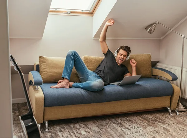 Hombre emocionado escuchando música en auriculares inalámbricos y gestos mientras está acostado en el sofá cerca de la computadora portátil - foto de stock