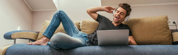 Imagen horizontal del hombre emocionado escuchando música en auriculares inalámbricos mientras está acostado en el sofá cerca de la computadora portátil - foto de stock