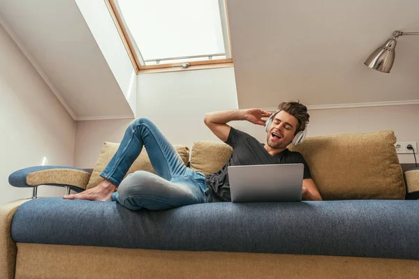 Homem animado ouvindo música em fones de ouvido sem fio enquanto deitado no sofá perto do laptop — Fotografia de Stock