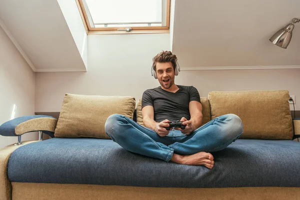 KIEW, UKRAINE - 13. APRIL 2019: Aufgeregter Mann mit drahtlosen Kopfhörern am Hals spielt Videospiel, während er mit gekreuzten Beinen auf dem Sofa sitzt — Stockfoto