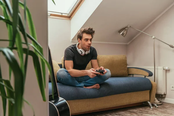 KYIV, UKRAINE - APRIL 13, 2019: selective focus of excited man in wireless headphones on neck playing video game while sitting on sofa — Stock Photo