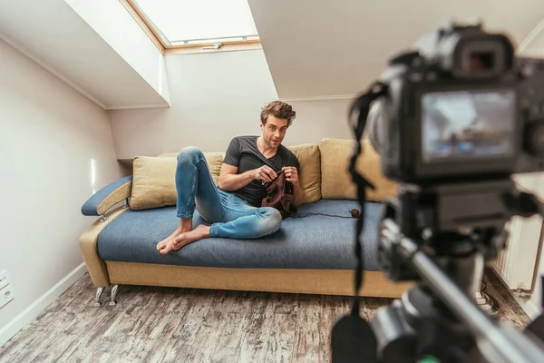 Selektiver Fokus junger Videoblogger, die auf dem Sofa stricken, während sie auf die Digitalkamera schauen — Stockfoto