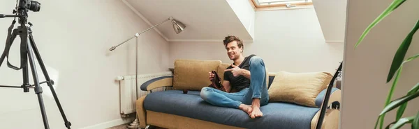 Handsome vlogger pointing with finger while knitting on sofa at home in attic room, horizontal image — Stock Photo
