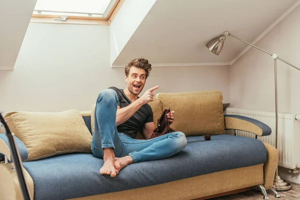 Aufgeregter junger Mann zeigt mit Finger beim Stricken auf Sofa zu Hause — Stockfoto