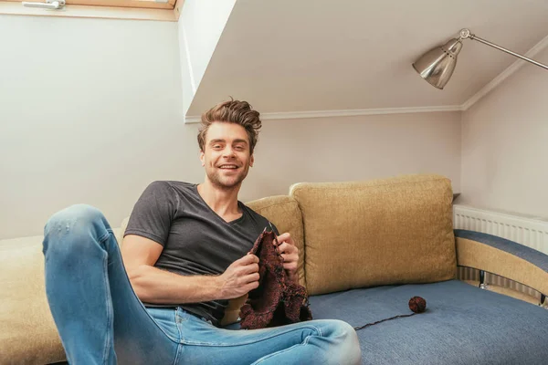 Glücklicher junger Mann schaut in die Kamera, während er zu Hause auf dem Sofa strickt — Stockfoto