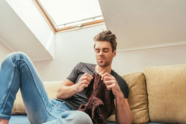 Low angle view of displeased man knitting while sitting on sofa in attic room — Stock Photo