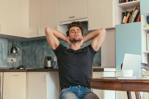 Erschöpfter Mann streckt sich mit Händen hinter dem Kopf, während er mit geschlossenen Augen in der Küche neben Laptop sitzt — Stockfoto