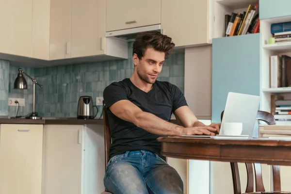 Hombre atento y sonriente escribiendo en el ordenador portátil en la cocina - foto de stock