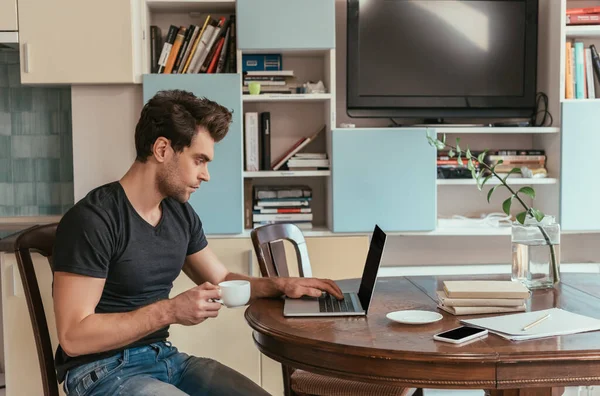 Vista lateral do homem pensativo segurando xícara de café enquanto trabalhava no laptop na cozinha — Fotografia de Stock