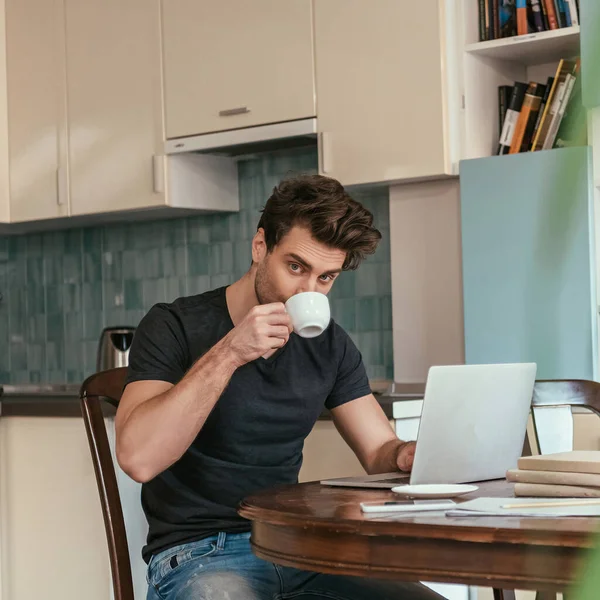 Hombre guapo bebiendo café y mirando a la cámara mientras trabaja en el ordenador portátil en la cocina — Stock Photo