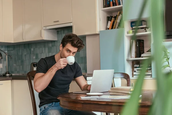 Foco seletivo de homem bonito bebendo café e olhando para a câmera enquanto trabalhava no laptop na cozinha — Fotografia de Stock