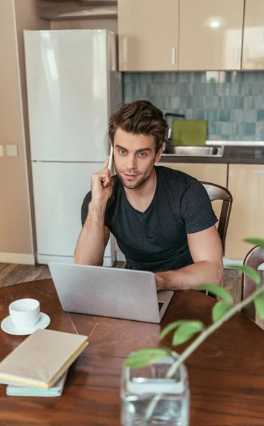 Selective focus of serious man talking on smartphone near laptop while working in kitchen — Stock Photo