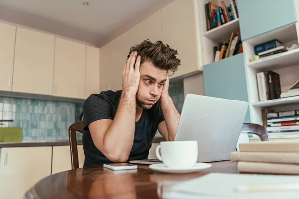 Hombre agotado tocando la cabeza mientras mira el monitor portátil cerca de la taza de café - foto de stock