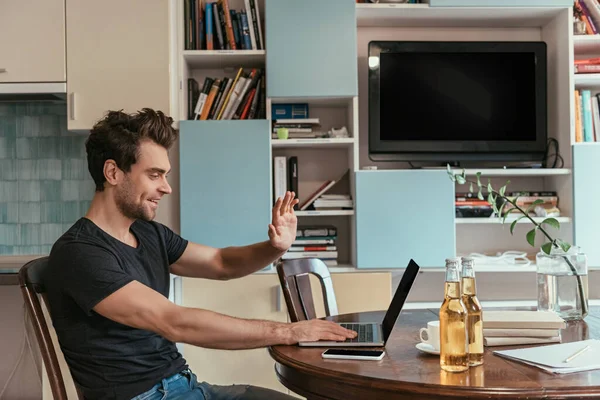 Vue latérale de l'homme joyeux agitant la main pendant le chat vidéo près des bouteilles de bière — Photo de stock