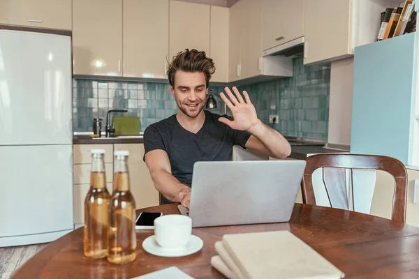 Alegre hombre saludando la mano durante el chat de vídeo en el portátil cerca de botellas de cerveza - foto de stock