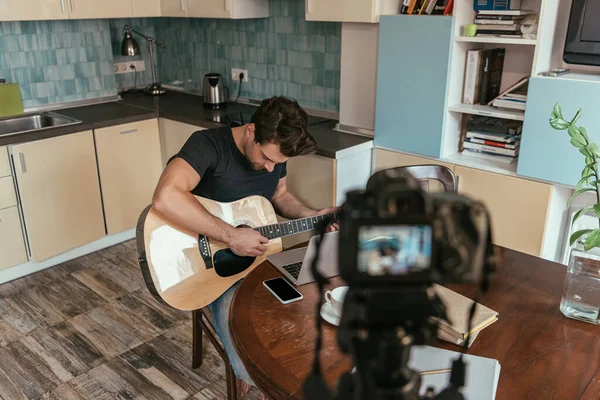 Vue grand angle du jeune vlogger jouant de la guitare dans la cuisine près de l'appareil photo numérique sur trépied — Photo de stock