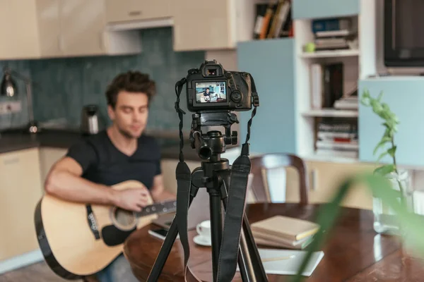 Selective focus of young vlogger playing guitar in kitchen near digital camera on tripod — Stock Photo