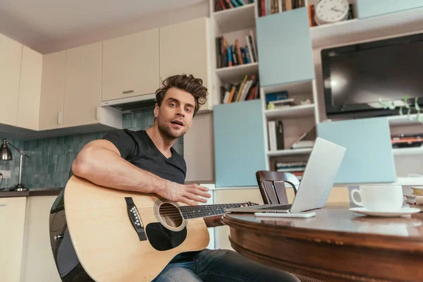 Vista de ángulo bajo del hombre guapo sosteniendo la guitarra mientras está sentado cerca de la computadora portátil - foto de stock