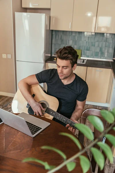 Foco seletivo de homem concentrado e bonito tocando guitarra enquanto olha para o laptop — Fotografia de Stock