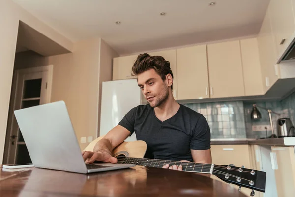 Beau jeune homme avec guitare en utilisant un ordinateur portable dans la cuisine — Photo de stock