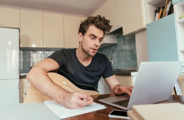 Concetrated young man with guitar holding pencil and using laptop — Stock Photo