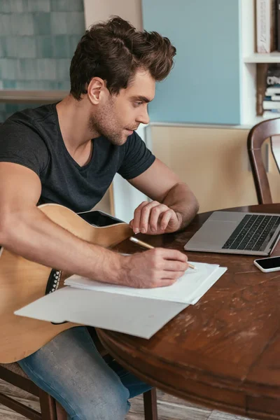 Konzentrierter junger Mann mit Gitarre, der Noten schreibt, während er auf Laptop schaut — Stockfoto