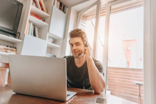 Ein lächelnder Mann blickt in die Kamera, während er zu Hause in der Nähe seines Laptops arbeitet — Stockfoto