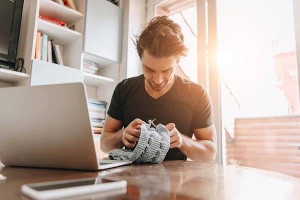 Attentive young man knitting while sitting near laptop at home — Stock Photo