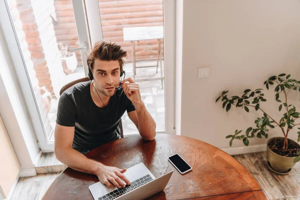 Vista de ángulo alto del hombre en auriculares que trabajan en casa cerca de la computadora portátil y el teléfono inteligente con pantalla en blanco - foto de stock