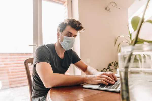Enfoque selectivo de hombre joven en máscara médica mirando a la cámara mientras trabaja en el ordenador portátil - foto de stock