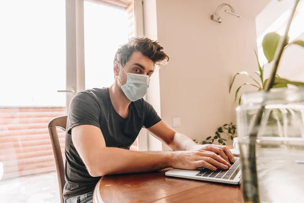 Foco seletivo do jovem na máscara médica olhando para a câmera enquanto trabalhava no laptop — Fotografia de Stock