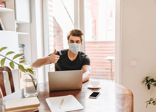 Junger Mann mit medizinischer Maske zeigt Daumen nach oben, während er zu Hause neben Laptop sitzt — Stockfoto