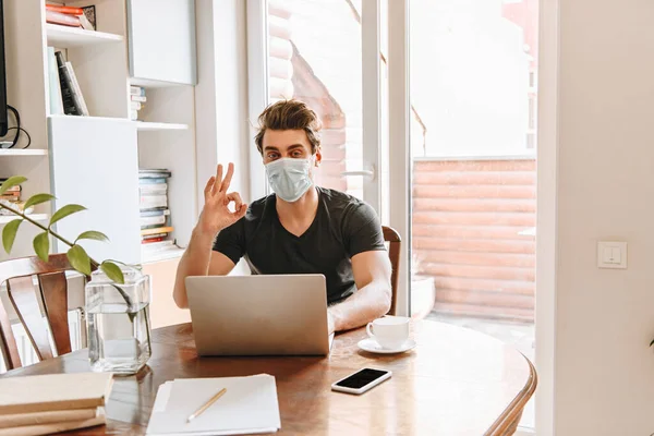 Joven en máscara protectora que muestra el pulgar hacia abajo cerca de la computadora portátil y la taza de café en la mesa — Stock Photo