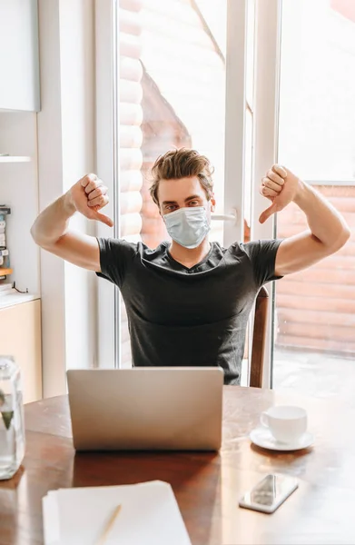 Giovane uomo in maschera medica mostrando pollici verso il basso vicino al computer portatile e una tazza di caffè sul tavolo — Foto stock