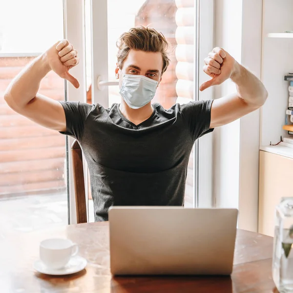 Joven en máscara médica que muestra los pulgares hacia abajo cerca de la computadora portátil y una taza de café - foto de stock