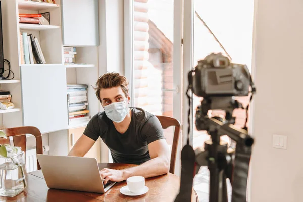 Selective focus of young vlogger in medical mask looking at digital camera while sitting near laptop — Stock Photo