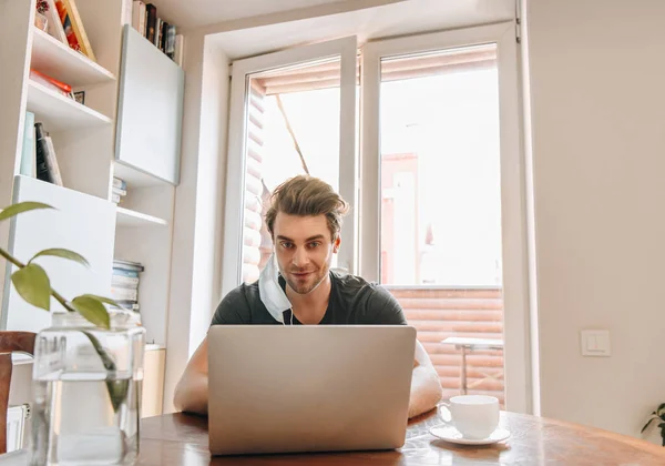 Homem sorridente com máscara médica pendurada na orelha trabalhando no laptop perto da xícara de café — Fotografia de Stock