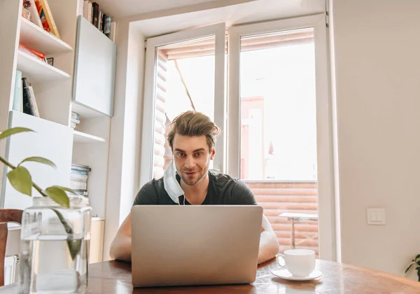 Lächelnder Mann mit medizinischer Maske am Ohr, der an einem Laptop neben einer Tasse Kaffee arbeitet — Stockfoto