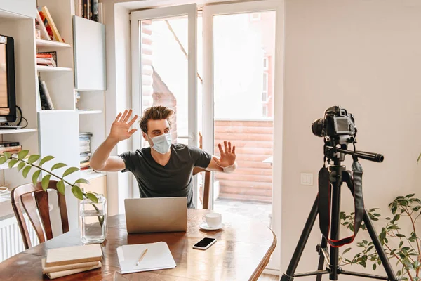 Joven video blogger en gestos de máscara médica mientras mira la cámara digital en el trípode - foto de stock