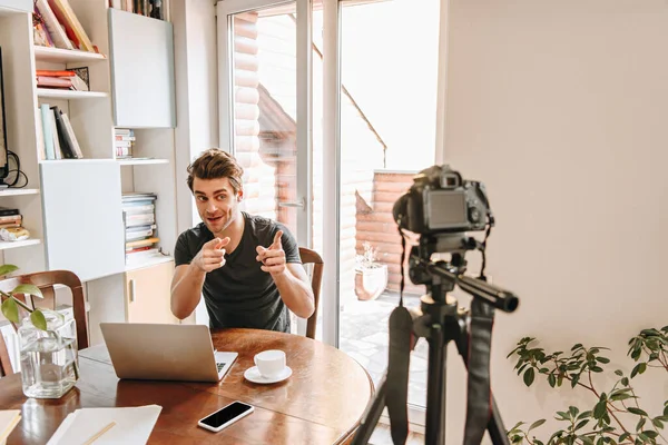 Веселый vlogger указывая пальцами на цифровую камеру, сидя рядом с ноутбуком — стоковое фото