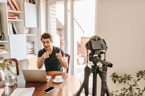 Alegre vlogger apuntando con los dedos a la cámara digital mientras está sentado cerca de la computadora portátil - foto de stock