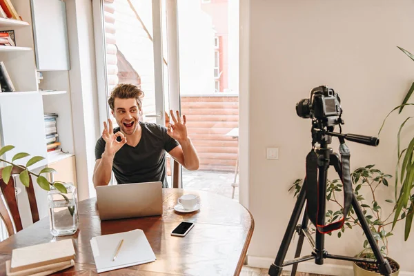 Веселий відеоблогер показує добре жести, сидячи біля ноутбука і дивлячись на цифрову камеру на тринозі — стокове фото