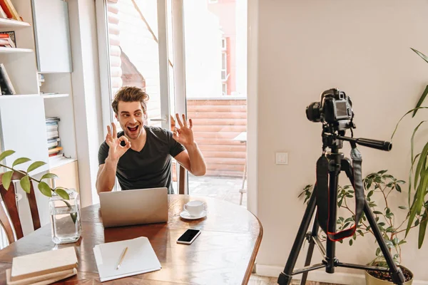 Alegre vlogger mostrando gestos bien mientras está sentado cerca de la computadora portátil y mirando a la cámara digital en el trípode - foto de stock