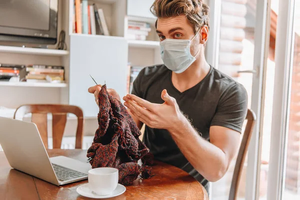 Junger Mann in medizinischer Maske sitzt mit offenen Armen in der Nähe von Laptop und Kaffeetasse und hält Strickfäden — Stockfoto