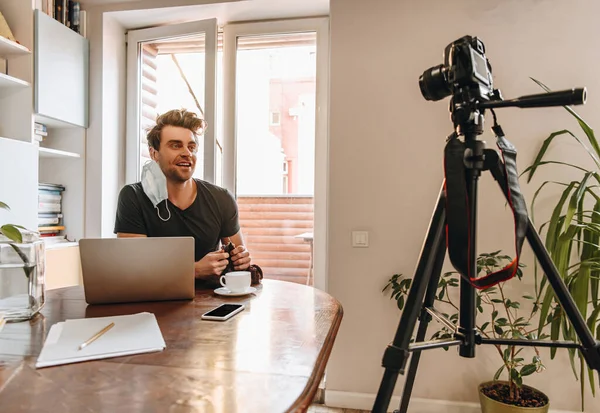 Smiling vlogger with medical mask on ear knitting while looking at digital camera on tripod — Stock Photo